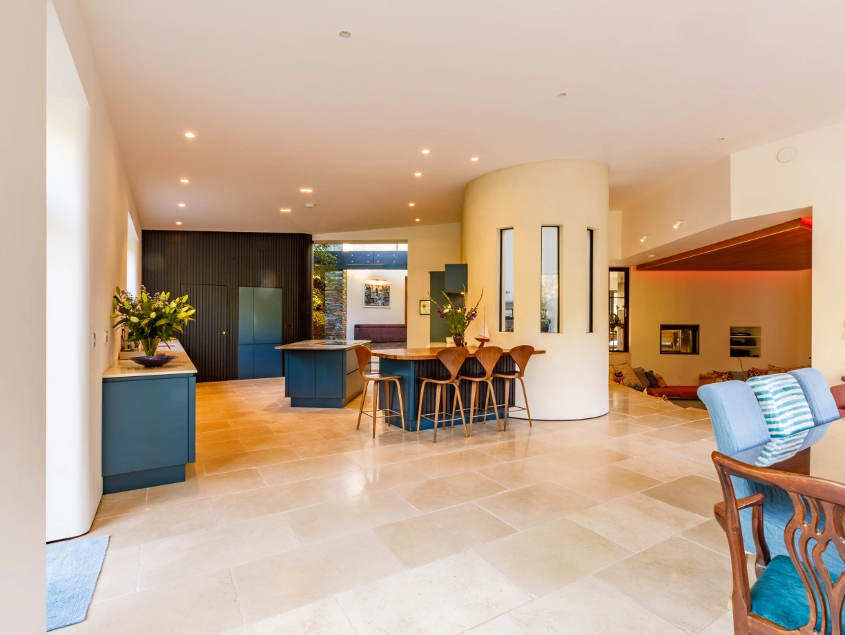 A spacious, modern kitchen and dining area featuring a large stone floor, light-colored walls, and contemporary furniture. The kitchen has dark cabinetry and a central island, while the dining space includes wooden chairs and a table. Natural light floods in through large windows.