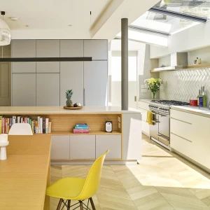 Cuisine moderne avec un plan de travail en bois et des armoires grises. Une table en bois avec des bougies blanches et une chaise jaune est au premier plan. Une fenêtre laisse entrer la lumière naturelle. Des plantes décoratives sont présentes.