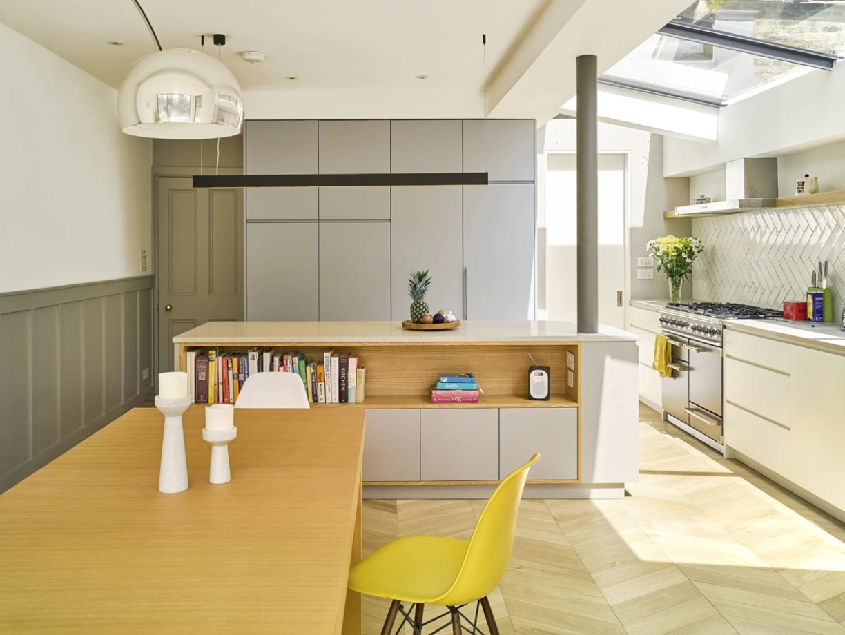 Cuisine moderne avec un plan de travail en bois et des armoires grises. Une table en bois avec des bougies blanches et une chaise jaune est au premier plan. Une fenêtre laisse entrer la lumière naturelle. Des plantes décoratives sont présentes.