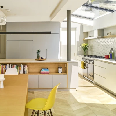 Cuisine moderne avec un plan de travail en bois et des armoires grises. Une table en bois avec des bougies blanches et une chaise jaune est au premier plan. Une fenêtre laisse entrer la lumière naturelle. Des plantes décoratives sont présentes.