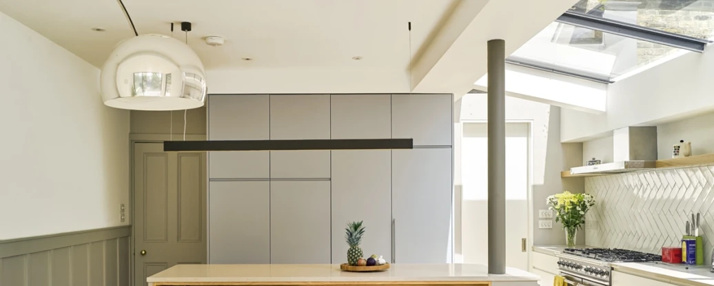 Cuisine moderne avec un plan de travail en bois et des armoires grises. Une table en bois avec des bougies blanches et une chaise jaune est au premier plan. Une fenêtre laisse entrer la lumière naturelle. Des plantes décoratives sont présentes.