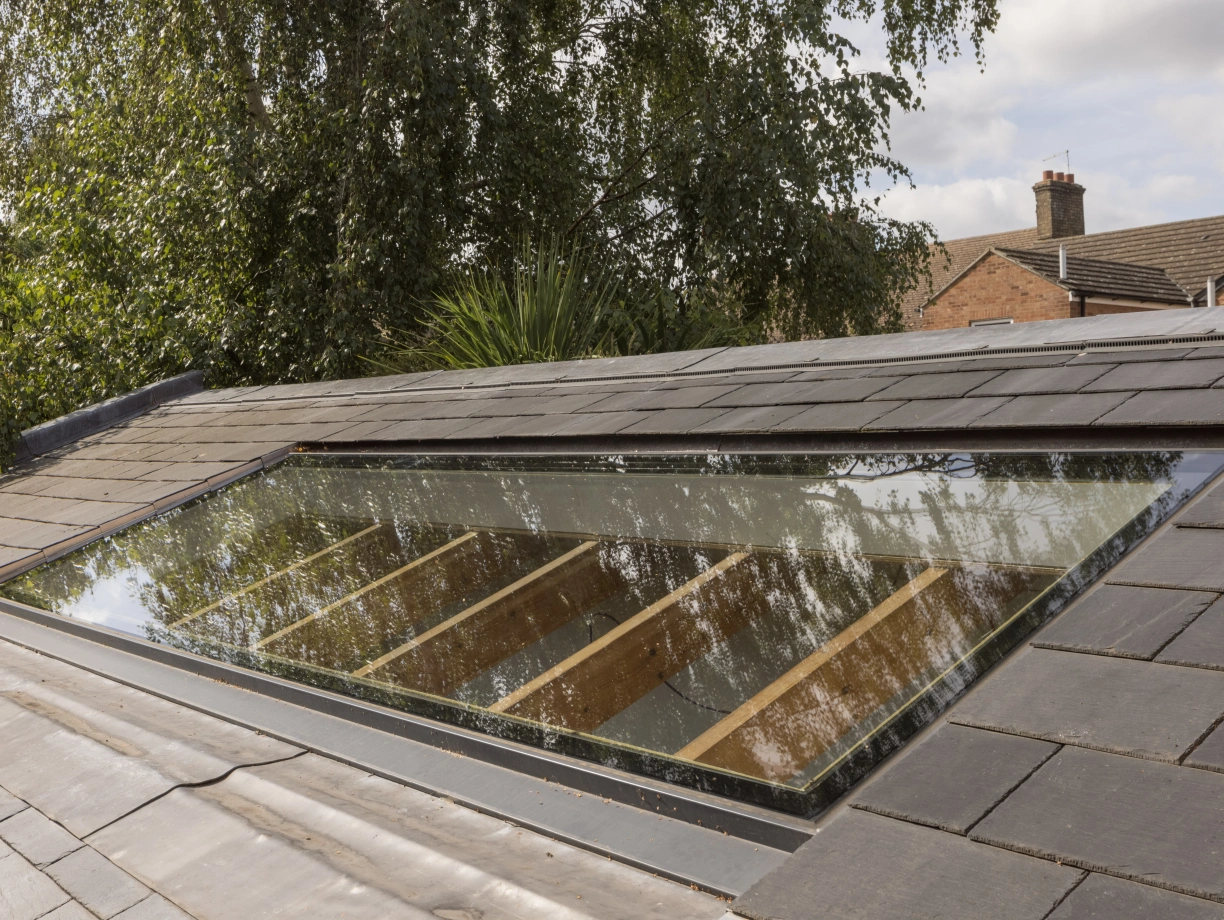 A skylight set into a slate roof, reflecting surrounding trees and buildings. Wooden beams can be seen through the glass of the skylight.