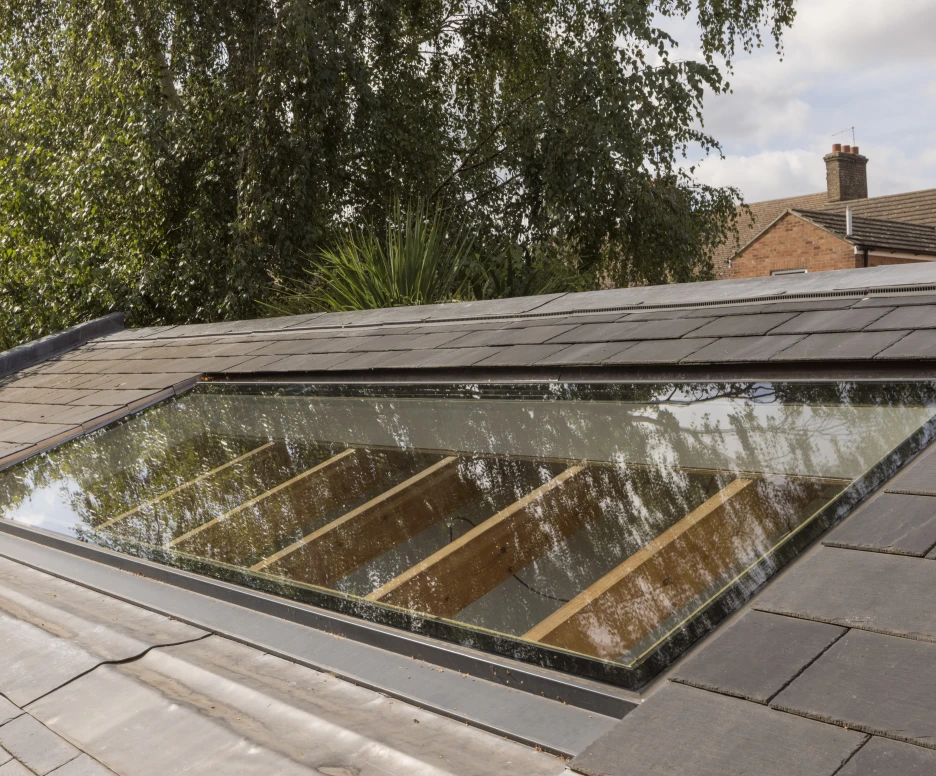 A skylight set into a slate roof, reflecting surrounding trees and buildings. Wooden beams can be seen through the glass of the skylight.