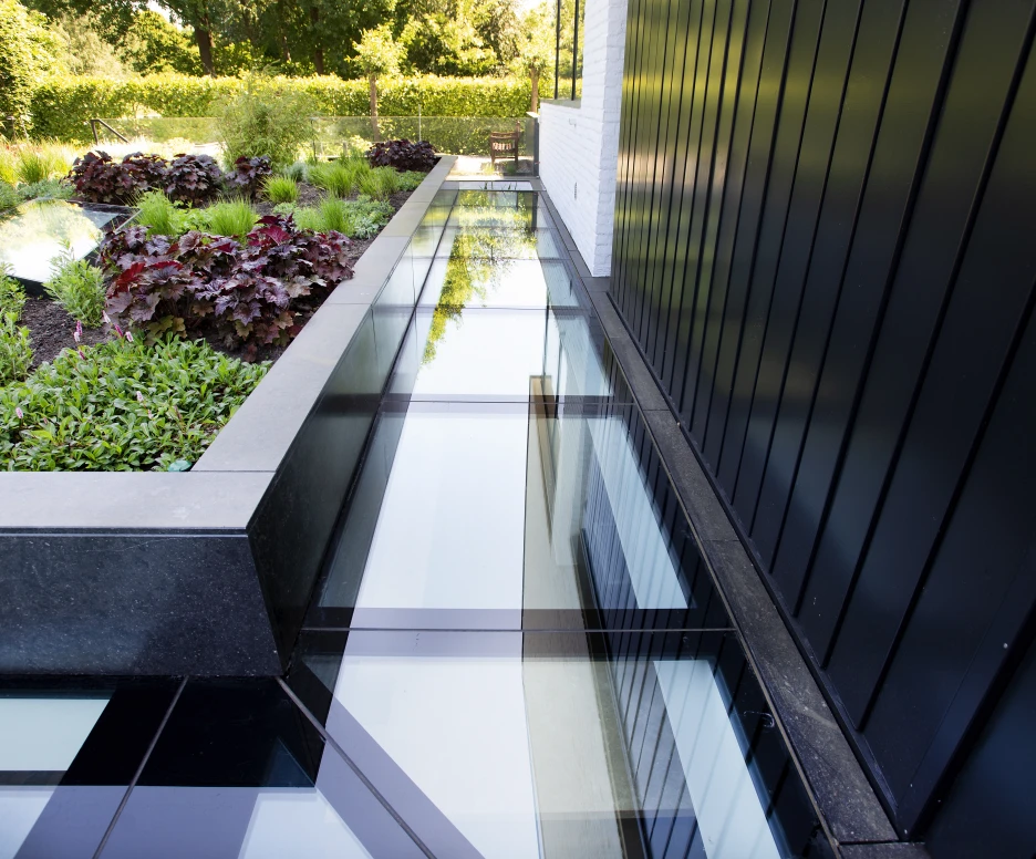 A modern outdoor space featuring glass walkways over a landscaped area with vibrant plants and dark stone borders. Lush greenery and colorful foliage provide a natural contrast to the sleek design.