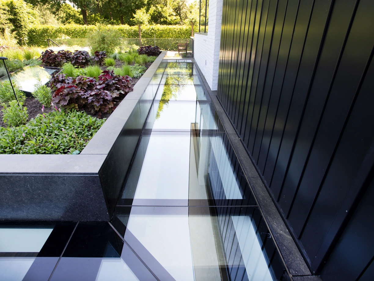 A modern outdoor space featuring glass walkways over a landscaped area with vibrant plants and dark stone borders. Lush greenery and colorful foliage provide a natural contrast to the sleek design.