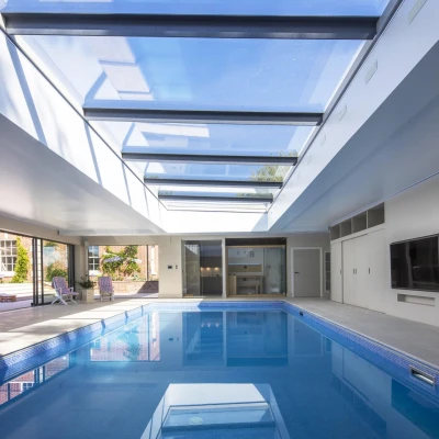An indoor swimming pool with a sleek design, featuring a glass ceiling that allows natural light to flood the space. Modern lounge chairs are positioned around the pool, and large sliding doors lead to an outdoor area.