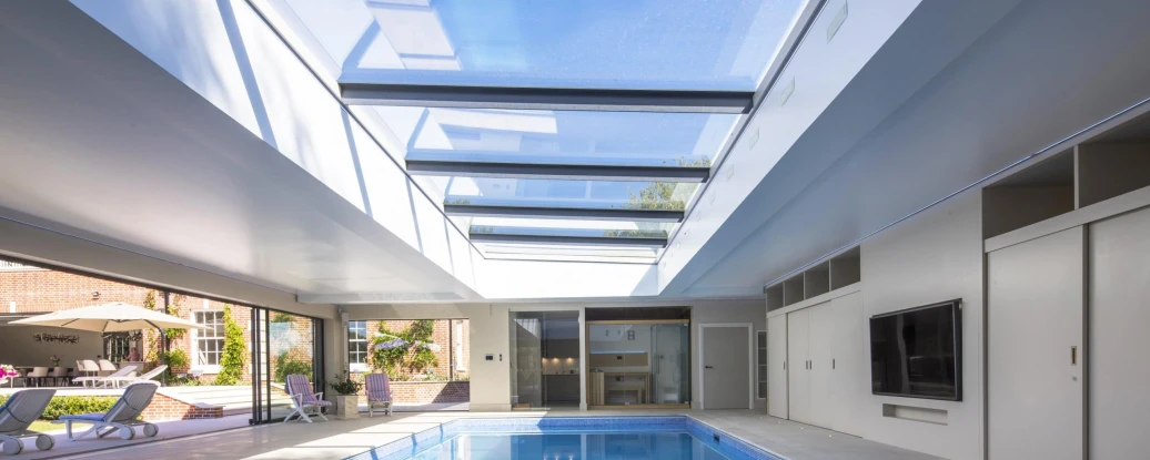 An indoor swimming pool with a sleek design, featuring a glass ceiling that allows natural light to flood the space. Modern lounge chairs are positioned around the pool, and large sliding doors lead to an outdoor area.