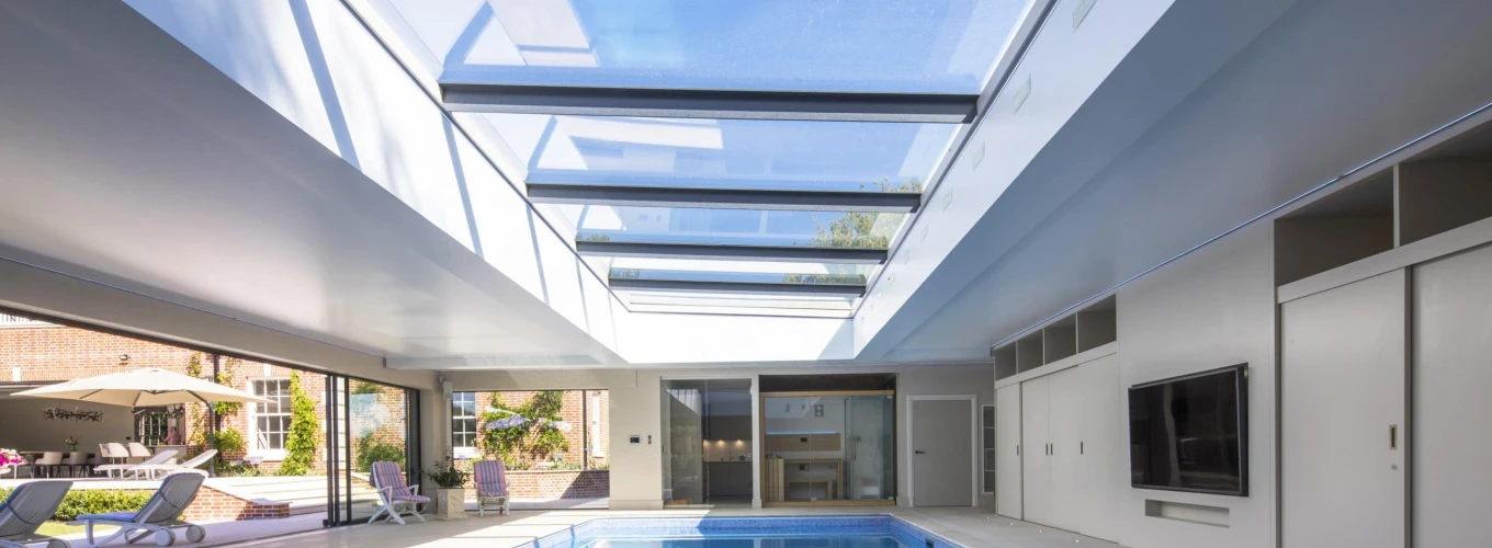 Intérieur d’une piscine avec un plafond en verre laissant passer la lumière naturelle. Des murs modernes et des portes coulissantes s’intègrent parfaitement au design. Des chaises longues sont disposées près de la piscine.