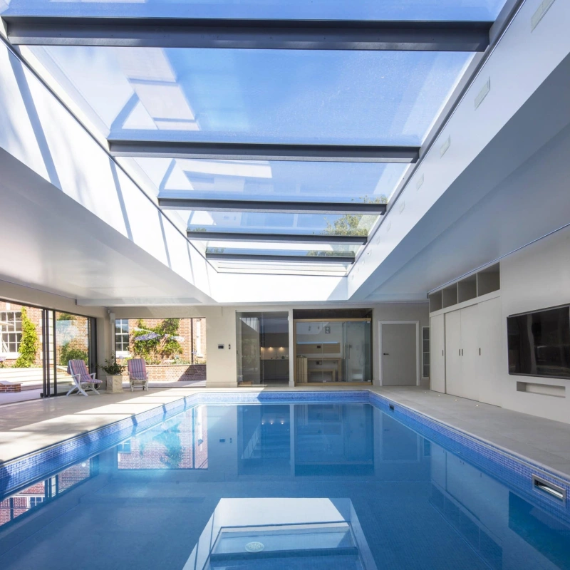 An indoor swimming pool with a sleek design, featuring a glass ceiling that allows natural light to flood the space. Modern lounge chairs are positioned around the pool, and large sliding doors lead to an outdoor area.