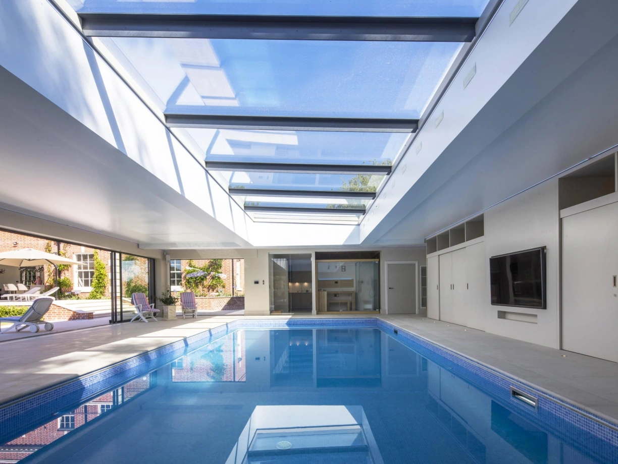Intérieur d’une piscine avec un plafond en verre laissant passer la lumière naturelle. Des murs modernes et des portes coulissantes s’intègrent parfaitement au design. Des chaises longues sont disposées près de la piscine.