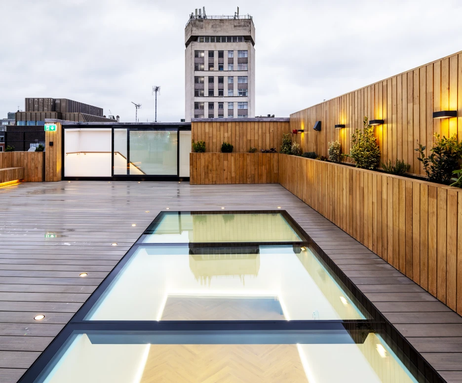 A modern rooftop terrace featuring a glass-bordered swimming pool reflecting the sky, surrounded by wooden decking and illuminated by soft lighting. In the background, a tall building with a classic facade is visible against a cloudy sky.