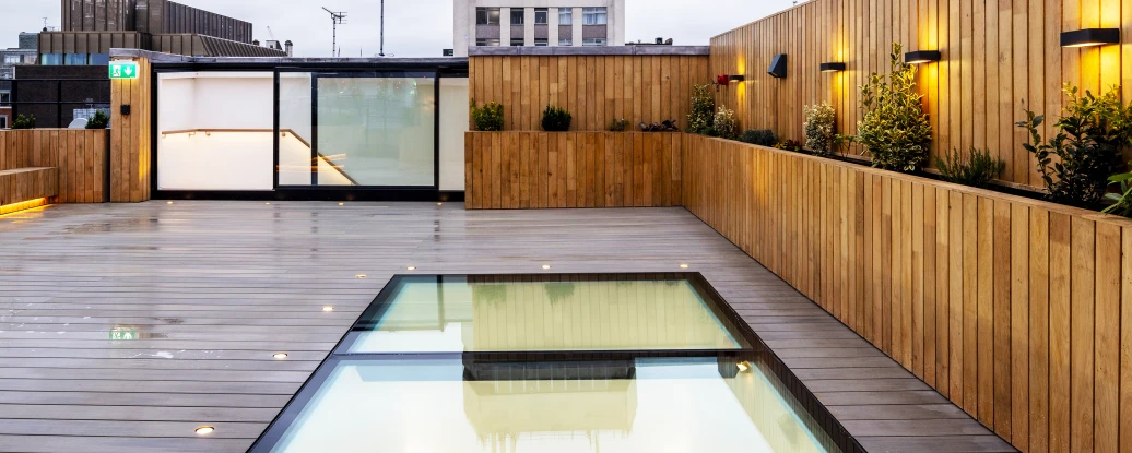 A modern rooftop terrace featuring a glass-bordered swimming pool reflecting the sky, surrounded by wooden decking and illuminated by soft lighting. In the background, a tall building with a classic facade is visible against a cloudy sky.