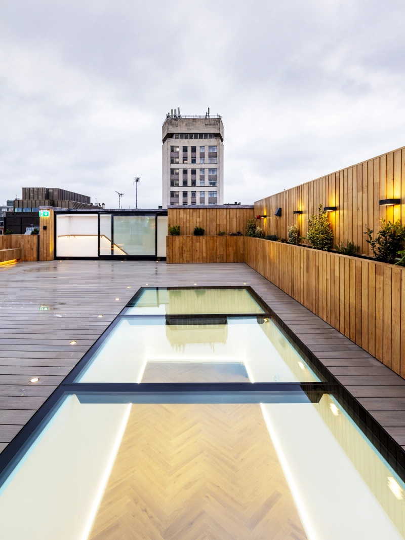 A modern rooftop with a wooden deck and a glass-lined swimming pool reflecting the overcast sky. A tall building stands in the background, adding to the urban atmosphere. Soft lighting accents the space, creating a warm ambiance.