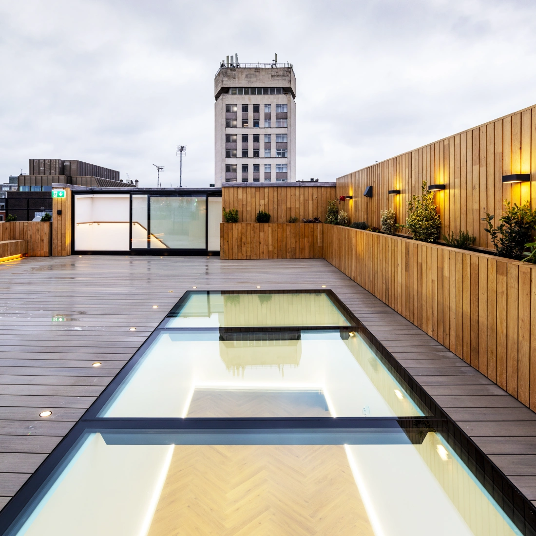 A modern rooftop terrace featuring a glass-bordered swimming pool reflecting the sky, surrounded by wooden decking and illuminated by soft lighting. In the background, a tall building with a classic facade is visible against a cloudy sky.