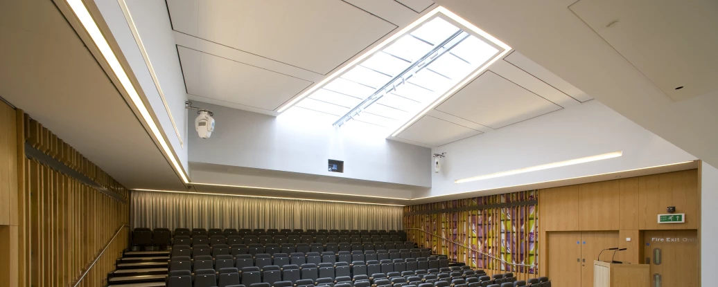 A modern auditorium with tiered black seating, wooden walls, and a large skylight. A wooden table with chairs is positioned at the front, and colorful artwork decorates one wall.