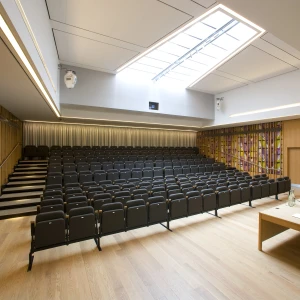 A modern auditorium with tiered black seating, wooden walls, and a large skylight. A wooden table with chairs is positioned at the front, and colorful artwork decorates one wall.