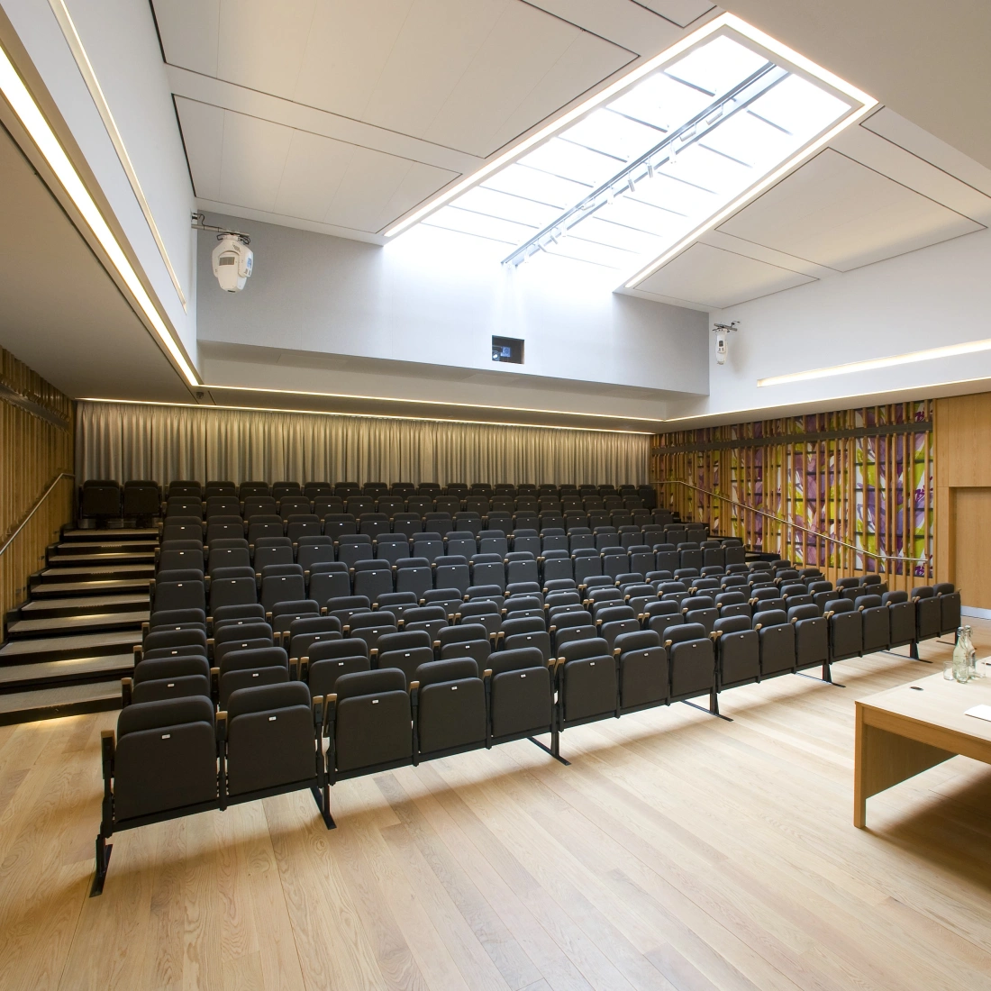 A modern auditorium featuring rows of black chairs, a wooden stage with a table and chairs, and large windows allowing natural light. The walls are adorned with wooden paneling and colorful stained glass.