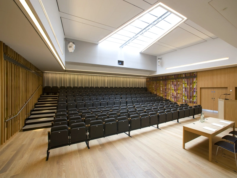 A modern auditorium featuring rows of black chairs, a wooden stage with a table and chairs, and large windows allowing natural light. The walls are adorned with wooden paneling and colorful stained glass.