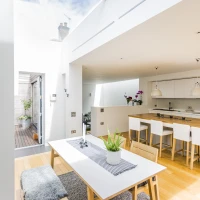 Luminoso interior de cocina moderna con mesa blanca, sillas de madera y plantas. Ventanas amplias permiten entrada de luz natural. Piso de madera clara.