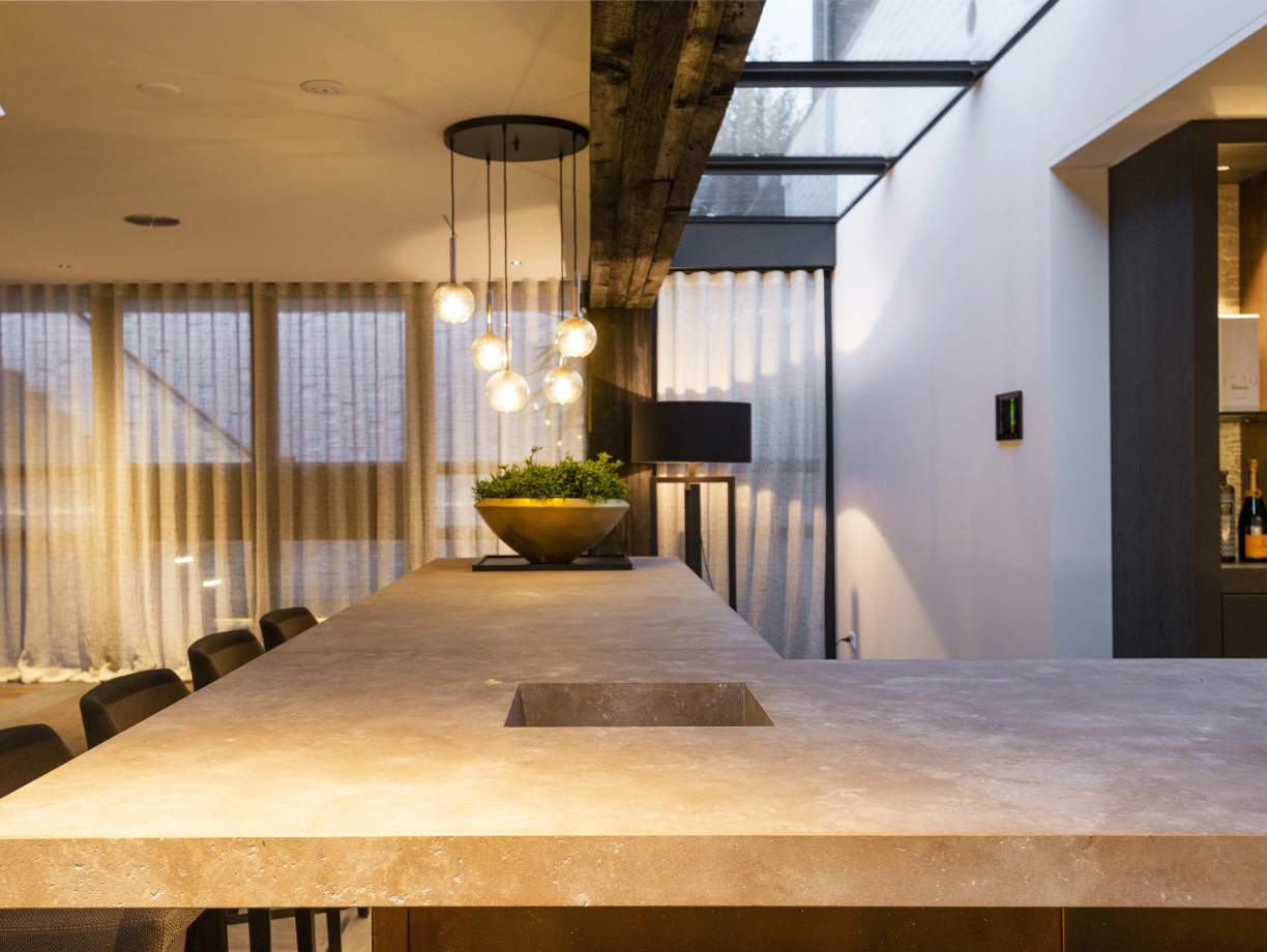 Cocina moderna con una barra de piedra, luces colgantes y una planta decorativa. Ventanas con cortinas suaves que permiten la entrada de luz.