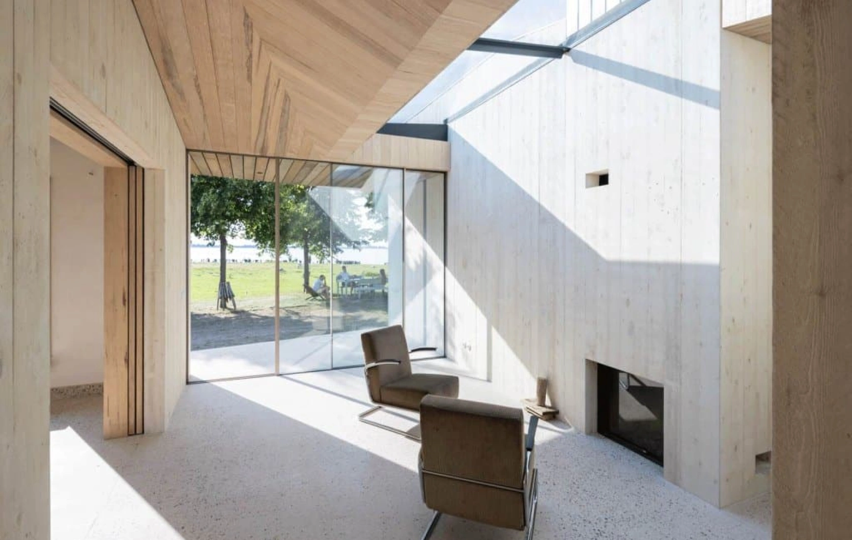 Intérieur d'une maison moderne avec des murs en bois clair et de grandes fenêtres laissant entrer la lumière naturelle. Une chaise en cuir grise est placée face à une cheminée. On aperçoit à l'extérieur une vue sur un espace vert.