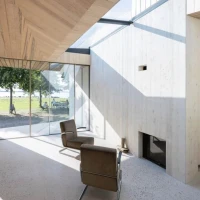 Heller Wohnraum mit Holzverkleidung, großen Fenstern und einem Blick auf die Landschaft. Ein moderner Stuhl steht im Raum, während Sonnenlicht durch die Fenster strömt.