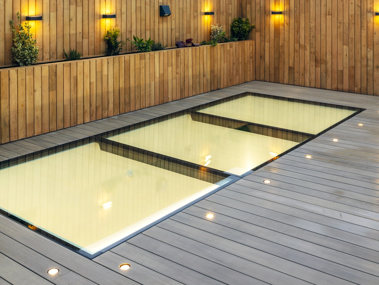 A rooftop space featuring a shallow pool surrounded by wooden decking and walls. Soft yellow lights illuminate the area, with small plants placed along the walls. A cloudy sky is visible in the background, along with a ferris wheel.