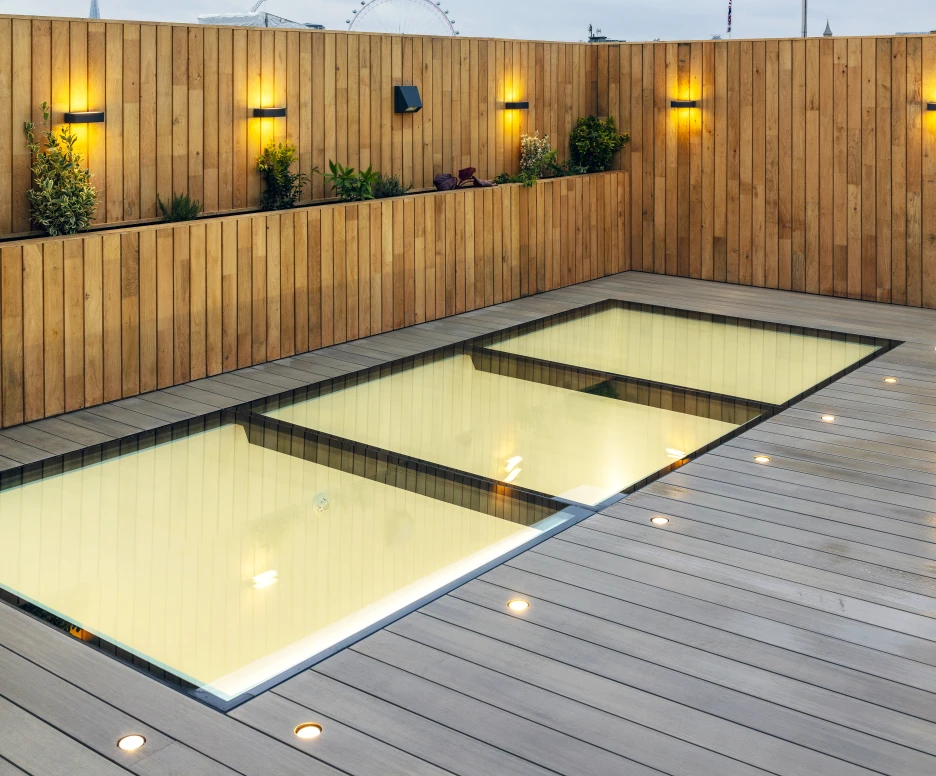 A rooftop space featuring a shallow pool surrounded by wooden decking and walls. Soft yellow lights illuminate the area, with small plants placed along the walls. A cloudy sky is visible in the background, along with a ferris wheel.