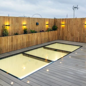 A sleek rooftop area featuring a modern, glass-walled pool surrounded by wooden decking. The wooden fence is adorned with small wall-mounted lights and plants. A cloudy sky can be seen in the background, along with a glimpse of a Ferris wheel.
