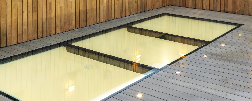 A sleek rooftop area featuring a modern, glass-walled pool surrounded by wooden decking. The wooden fence is adorned with small wall-mounted lights and plants. A cloudy sky can be seen in the background, along with a glimpse of a Ferris wheel.
