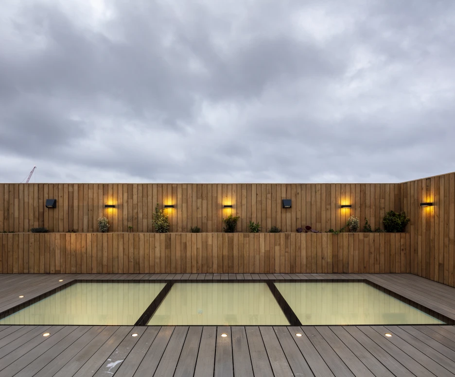 A modern rooftop terrace features two glass-paneled areas revealing a pool beneath. Wooden walls adorned with wall-mounted lights and small plants create a cozy ambiance. Overcast skies loom above, adding a tranquil mood to the scene.