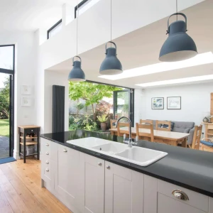 Modern kitchen showcasing a black countertop, white cabinets, and two pendant lights. Large glass doors lead to a backyard with patio furniture and greenery. A dining area is visible in the background.