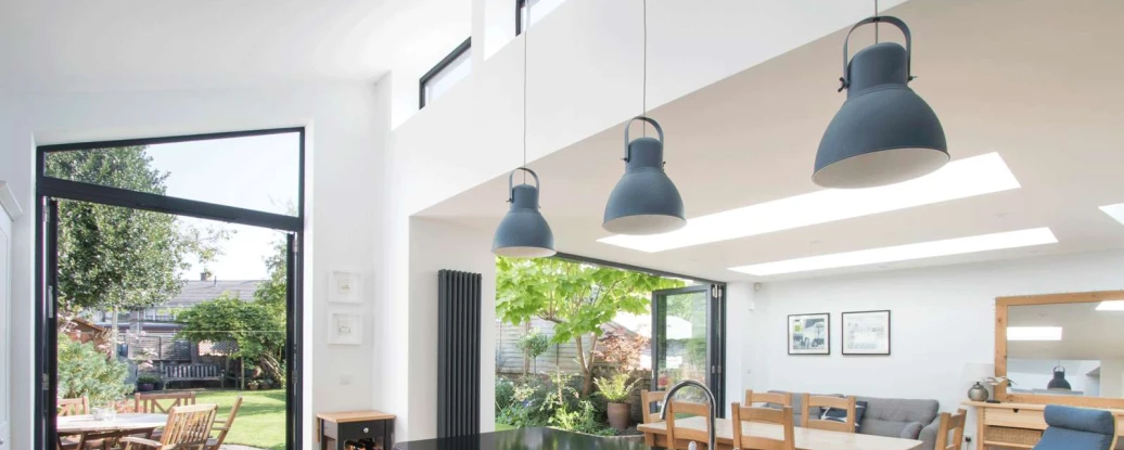 Modern kitchen showcasing a black countertop, white cabinets, and two pendant lights. Large glass doors lead to a backyard with patio furniture and greenery. A dining area is visible in the background.