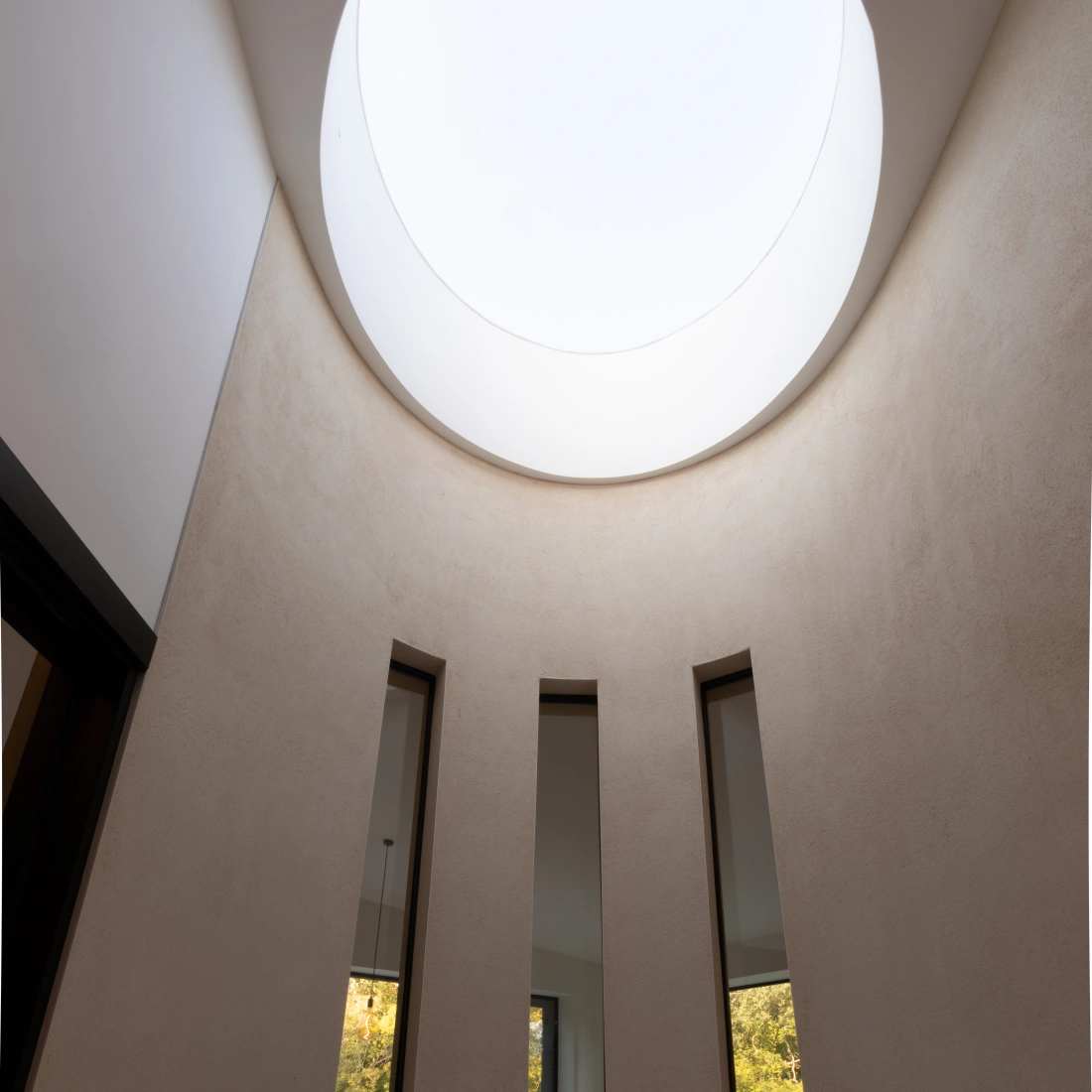 A modern interior space with smooth, beige walls and three tall, narrow windows. A circular skylight above allows natural light to illuminate the room. The floor is flat and minimalistic, enhancing the airy atmosphere.