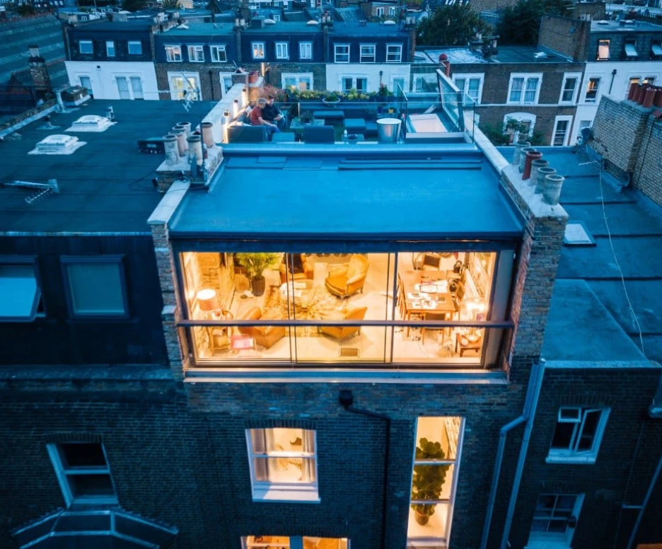 Aerial view of a modern building showcasing a rooftop terrace and large glass windows, revealing a cozy interior with seating and plants. The surrounding rooftops are visible in the background during twilight.