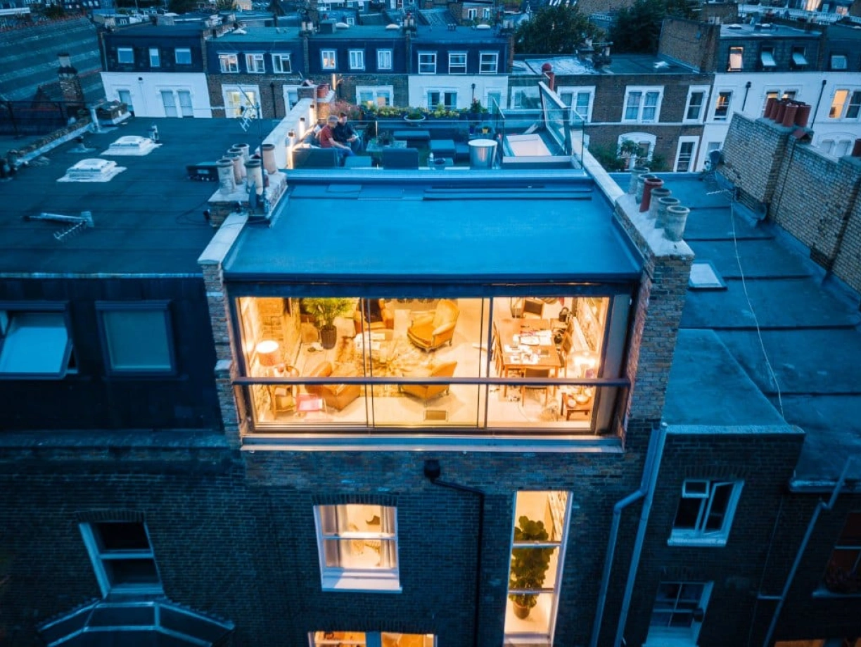Aerial view of a modern building showcasing a rooftop terrace and large glass windows, revealing a cozy interior with seating and plants. The surrounding rooftops are visible in the background during twilight.