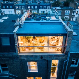 Aerial view of a modern building showcasing a rooftop terrace and large glass windows, revealing a cozy interior with seating and plants. The surrounding rooftops are visible in the background during twilight.