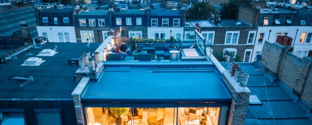 A modern rooftop terrace with a glass-walled lounge area, featuring cozy furniture and decorative plants. Nearby rooftops of city buildings are visible in the background, illuminated by evening lights.