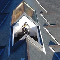 Ventana en el techo de una casa con forma triangular, que muestra el interior. Hay un tambor y un objeto negro en el espacio iluminado.