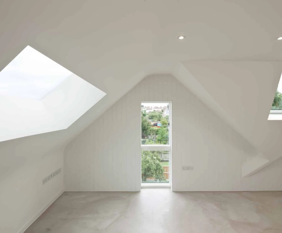 A minimalist room with slanted white walls, featuring two distinct windows that provide natural light and a view of greenery outside. The floor is light-colored and the space appears spacious and modern.
