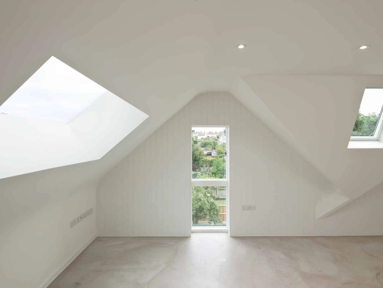 A minimalist room with slanted white walls, featuring two distinct windows that provide natural light and a view of greenery outside. The floor is light-colored and the space appears spacious and modern.