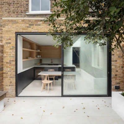 A modern kitchen view through glass doors, featuring minimalist design elements with wooden shelves, a white table, and two wooden chairs. The exterior showcases brick walls and a landscaped area with plants.