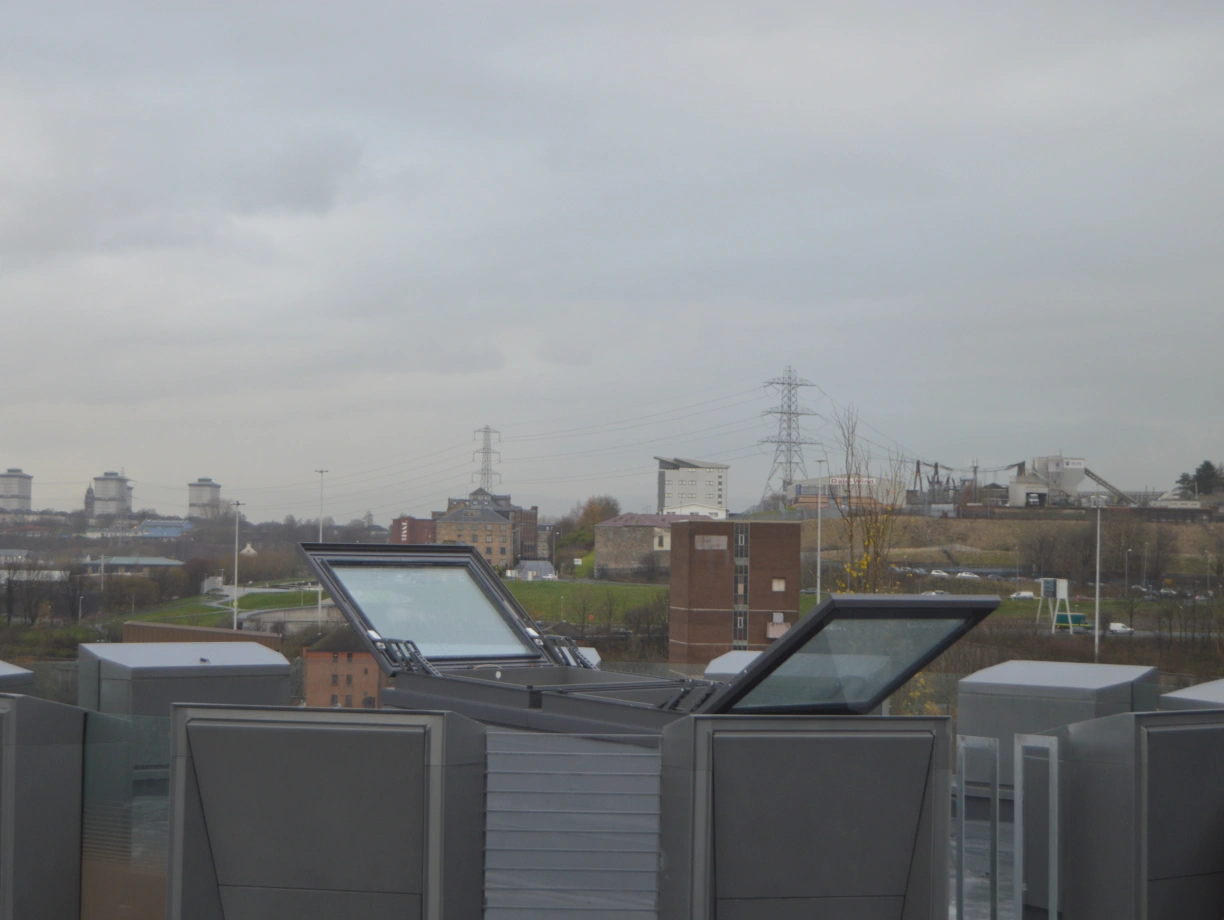 A view from a rooftop featuring several opened skylights, with a cloudy sky in the background. In the distance, a landscape includes buildings and power lines.