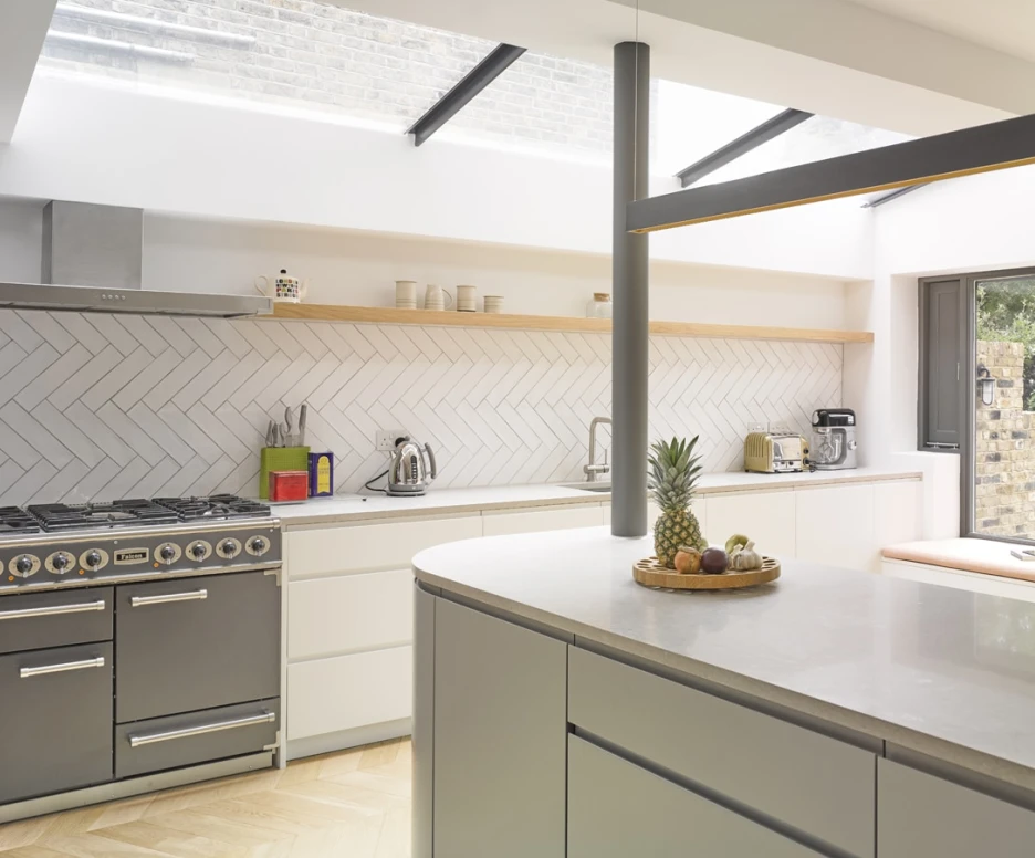 Cuisine moderne avec des meubles blancs, un plan de travail en pierre et un sol en bois clair. Un réfrigérateur, une bouilloire et une machine à café sont visibles sur le plan de travail. Une fenêtre apporte de la lumière naturelle. Une assiette de fruits est placée sur le comptoir.