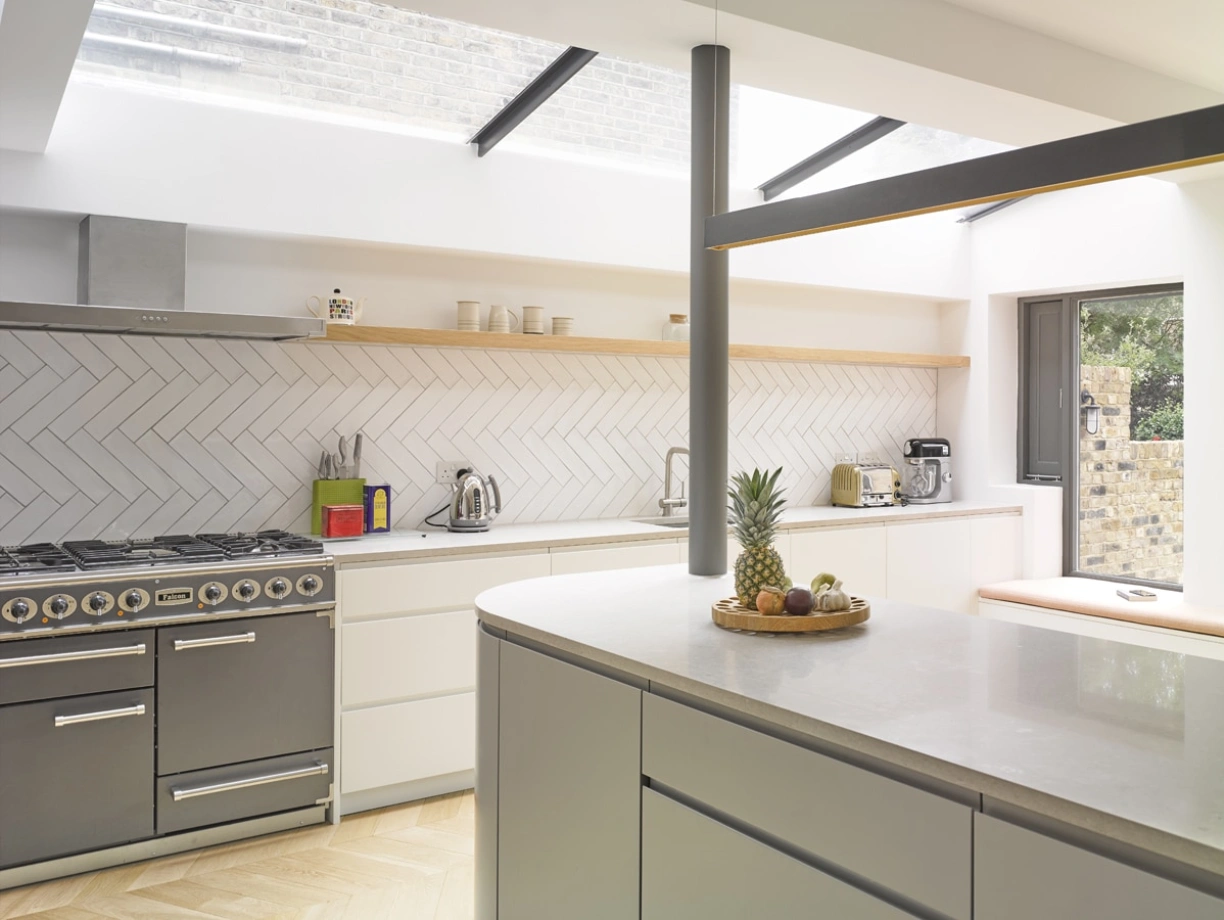 Cuisine moderne avec des meubles blancs, un plan de travail en pierre et un sol en bois clair. Un réfrigérateur, une bouilloire et une machine à café sont visibles sur le plan de travail. Une fenêtre apporte de la lumière naturelle. Une assiette de fruits est placée sur le comptoir.