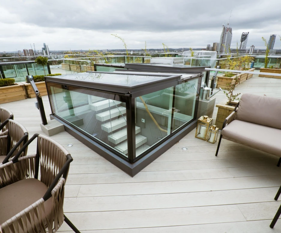A modern rooftop terrace features a glass enclosure and a staircase leading down. Stylish outdoor seating with a table is provided, along with planters and greenery. The skyline and tall buildings are visible in the background under a cloudy sky.