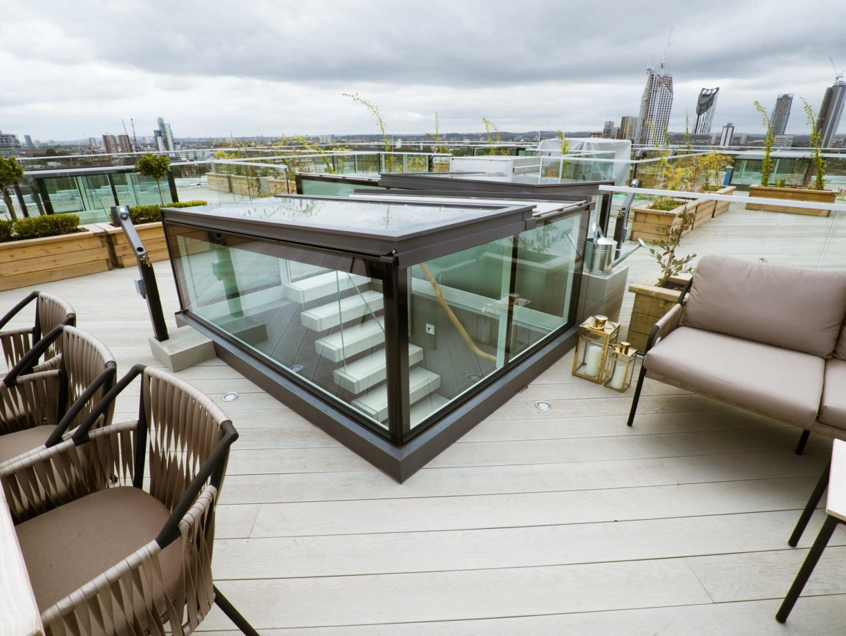A modern rooftop terrace features a glass enclosure and a staircase leading down. Stylish outdoor seating with a table is provided, along with planters and greenery. The skyline and tall buildings are visible in the background under a cloudy sky.