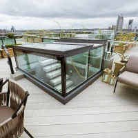 A rooftop terrace featuring modern glass stairs leading down, wooden planters with lush greenery, stylish outdoor seating, and a cloudy skyline in the background.