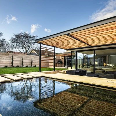 A modern house with a flat roof and wooden slatted overhang, surrounded by a manicured garden and a reflective pool. The design features large glass windows, offering a view of the outdoor space, which includes neatly trimmed shrubs and a landscaped lawn.