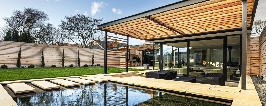 A modern house with a flat roof and wooden slatted overhang, surrounded by a manicured garden and a reflective pool. The design features large glass windows, offering a view of the outdoor space, which includes neatly trimmed shrubs and a landscaped lawn.